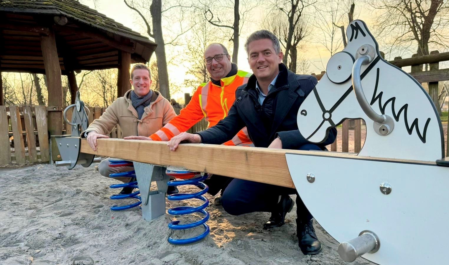 Drei Männer knien hinter einer Kinderwippe mit Pferdeköpfen vor den Sitzen.