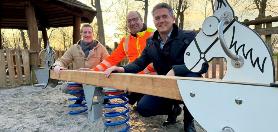 Drei Männer knien hinter einer Kinderwippe mit Pferdeköpfen vor den Sitzen.