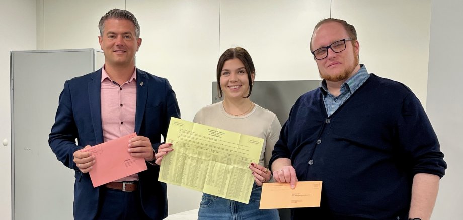 Drei Personen hinter einer Wahlurne. In den Händen halten sie Stimmzettel und farbige Briefwahlumschläge.