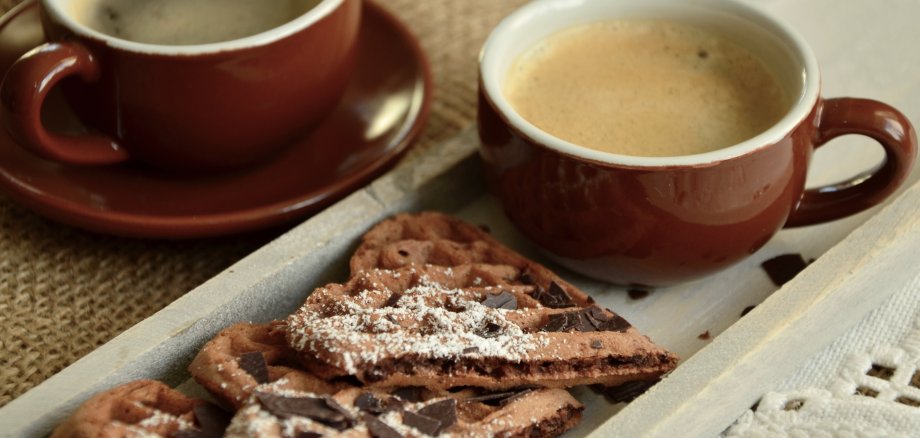 Auf einem Teller liegen mit Puderzucker bestäubte Waffelherzen. Zwei rote Kaffeetassen stehen drumherum.