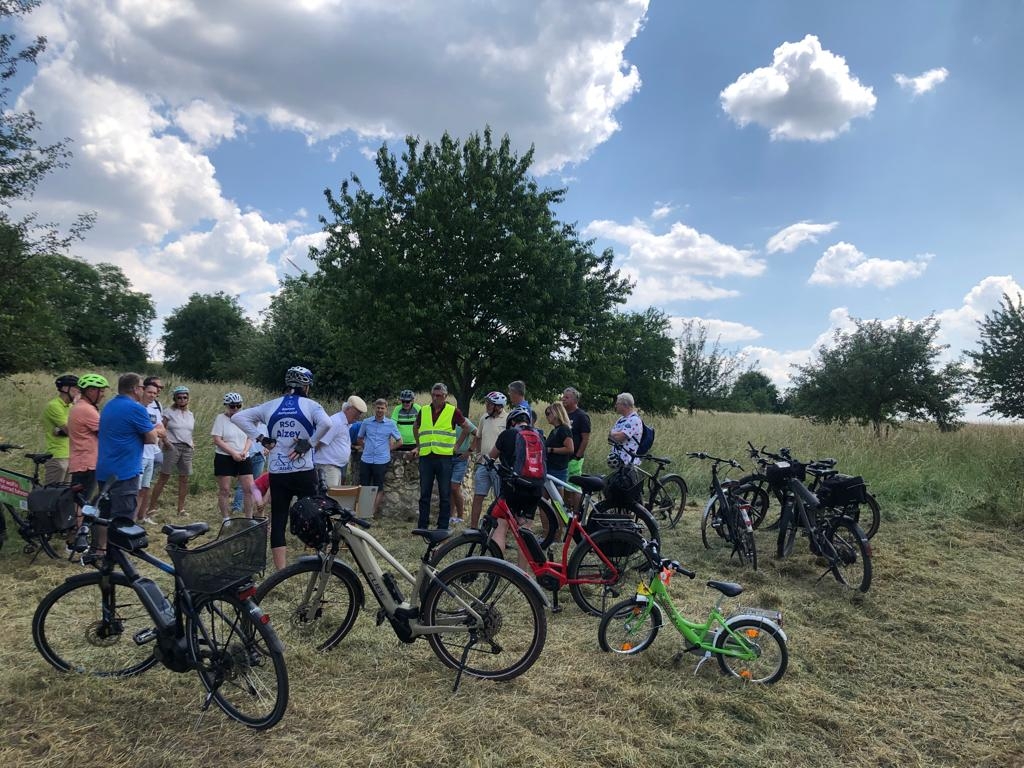 Eine Gruppe von Radfahrern macht in der grünen Landschaft Pause und hören einen Vortrag.