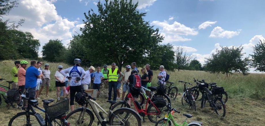 Eine Gruppe von Radfahrern macht in der grünen Landschaft Pause und hören einen Vortrag.