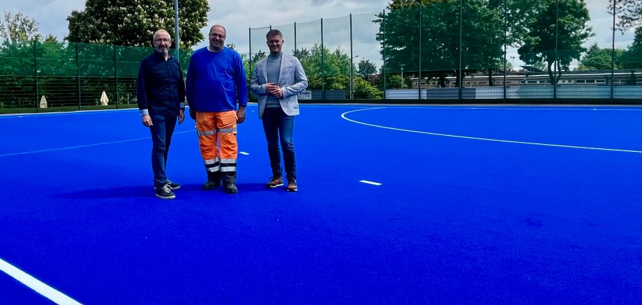 Drei Männer stehen auf einem strahlend blauen Hockeyrasen