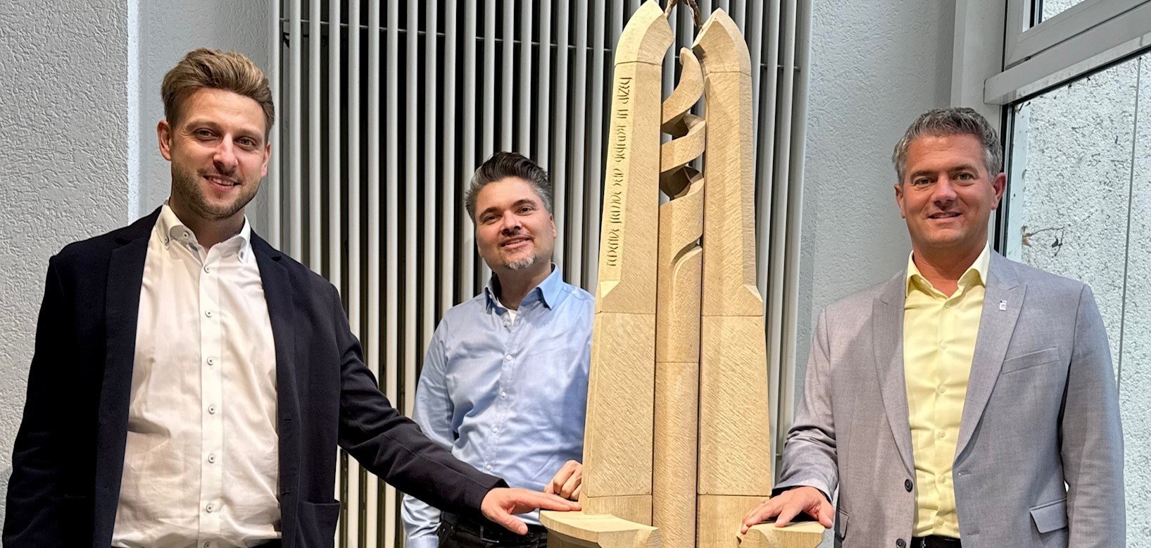 Drei Männer stehen um ein Kunstwerk aus Sandstein auf einem dunklen Würfel.