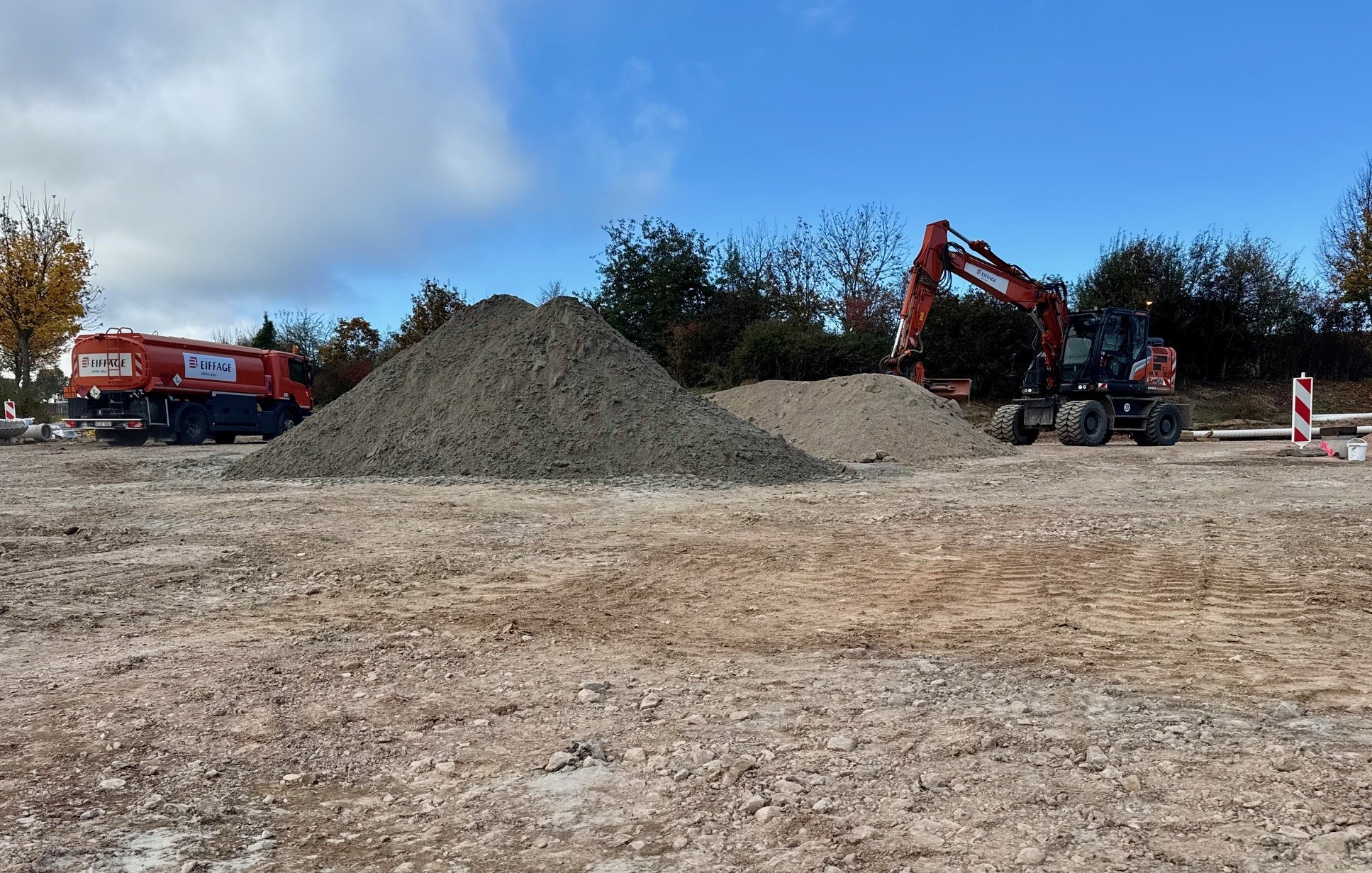Eine große Schotterfläche mit einem großen Sandberg. Bagger und Lastwagen fahren im Hintergrund.