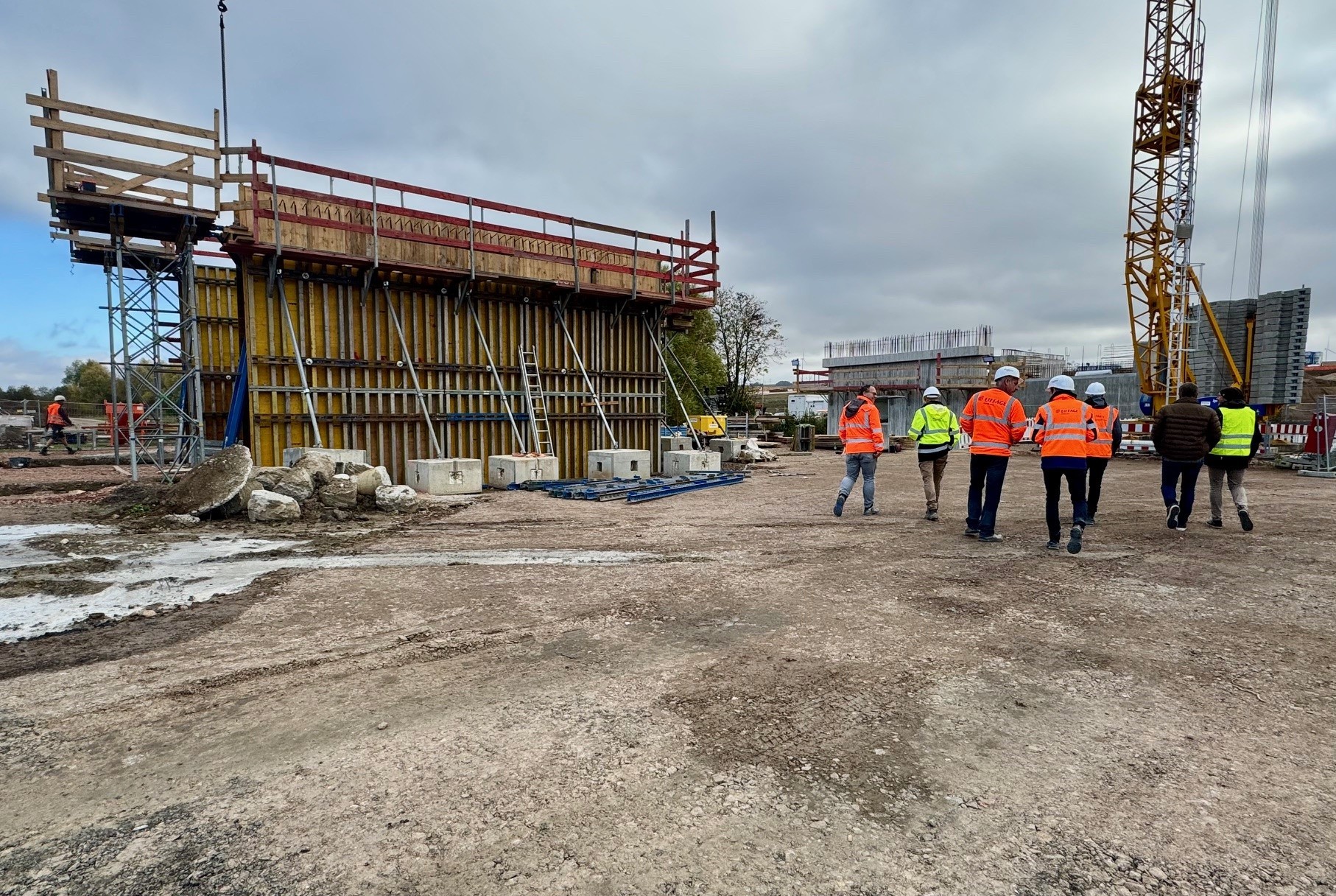 Ein großes Brückenteil auf einer großen Schotterfläche. Davor steht eine Gruppe von Menschen in bunten Jacken.
