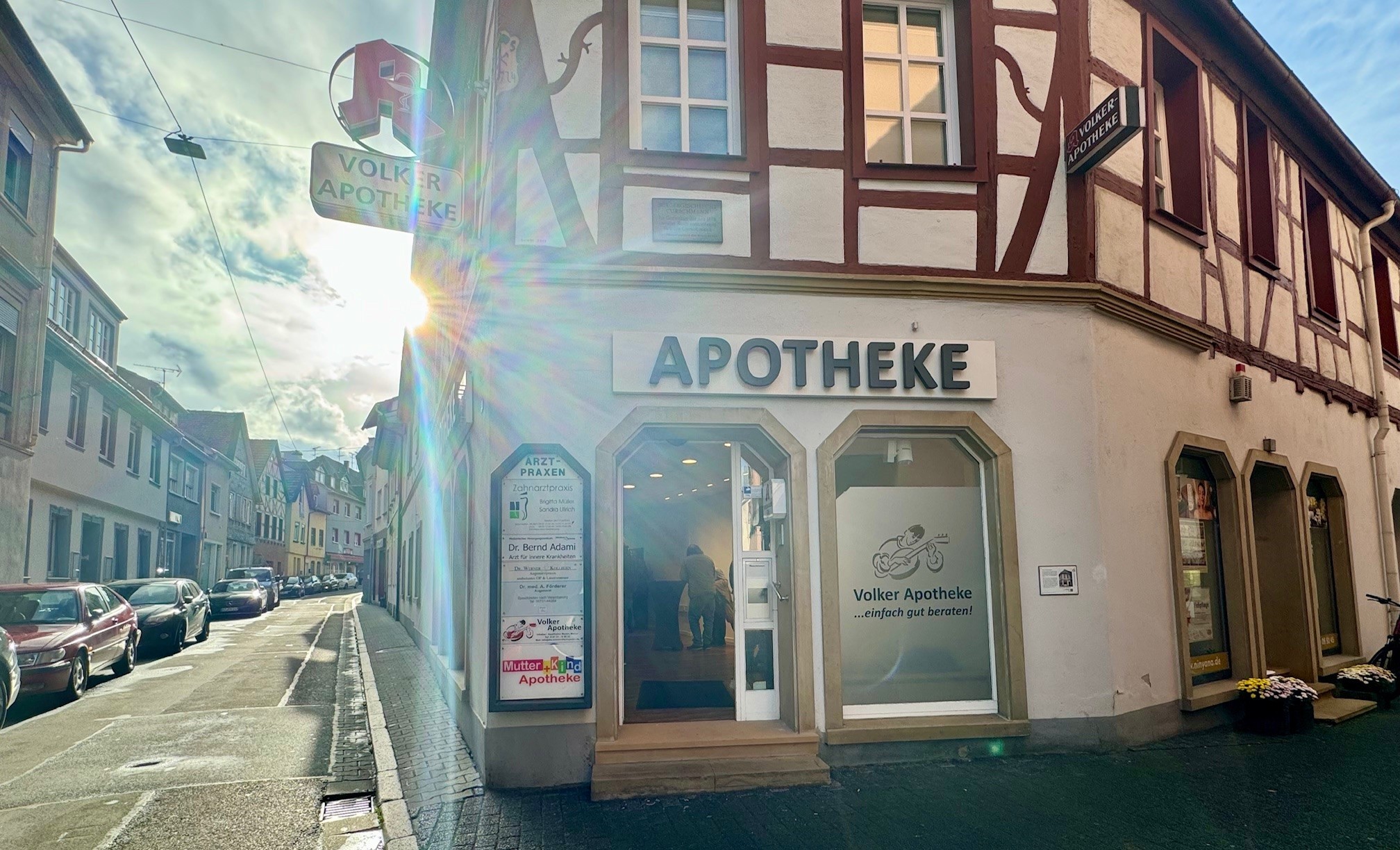Ein Fachwerkhaus. Über dem Gebäude steht Apotheke. Im Innern stehen Leute an Stehtischen.