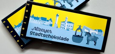 Eine Schokoladentafel in hellblau und gelbe mit Alzeyer Motiven im Hintergrund. 