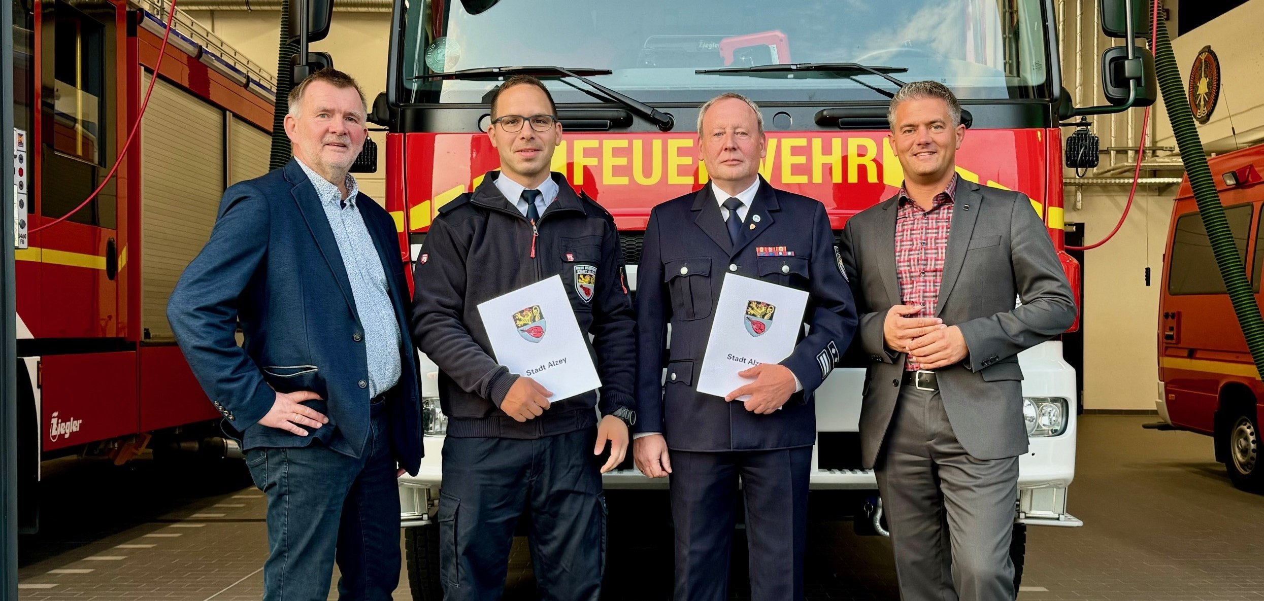 Vier Männer stehen vor einem Feuerwehrauto. Zwei von ihnen halten Urkunden in der Hand.