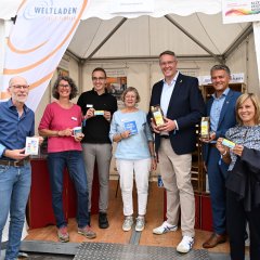 Man sieht eine kleine Gruppe von Menschen unter anderen auch Bürgermeister Steffen Jung vor einem Stand des Weltenladens Alzey e.V. die einige Fair Traid Produkte der Stadt Alzey halten unter anderem Kaffebohnen, Tassen, Schokolade mit dem Alzeyer Stadtlogo.