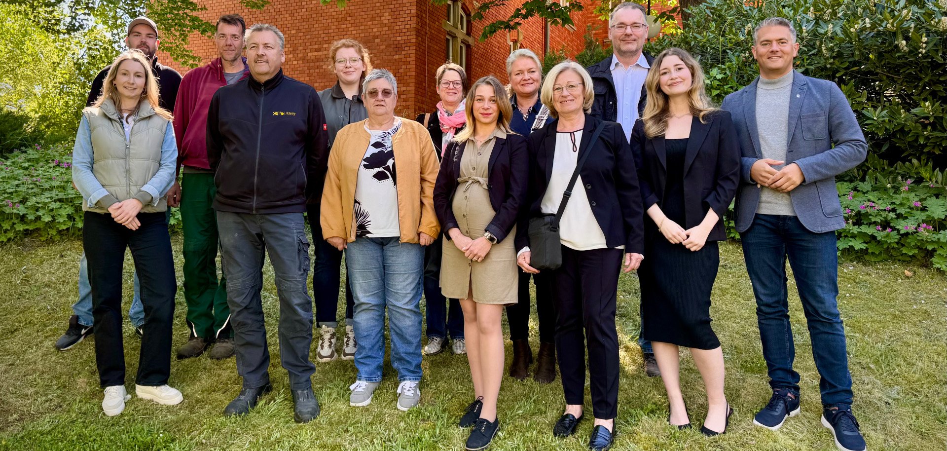 13 Personen stehen auf einer grünen Wiese hinter dem Rathaus