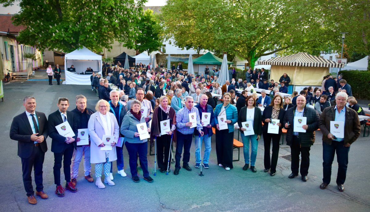 viele Personen im Hintergrund am sitzen/stehen/laufen und vorne eine Gruppe die weißes Blatt von Stadt Alzey halten