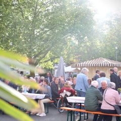 viele Personen sitzen an Holztischen und auf Sitzbänken und einige stehen/laufen