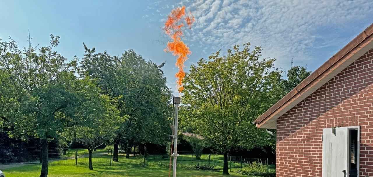 Gas wird in Form von einer Flamme abgefackelt im Hintergrund viele grüne Fläche und ein Haus mit Steinoptik