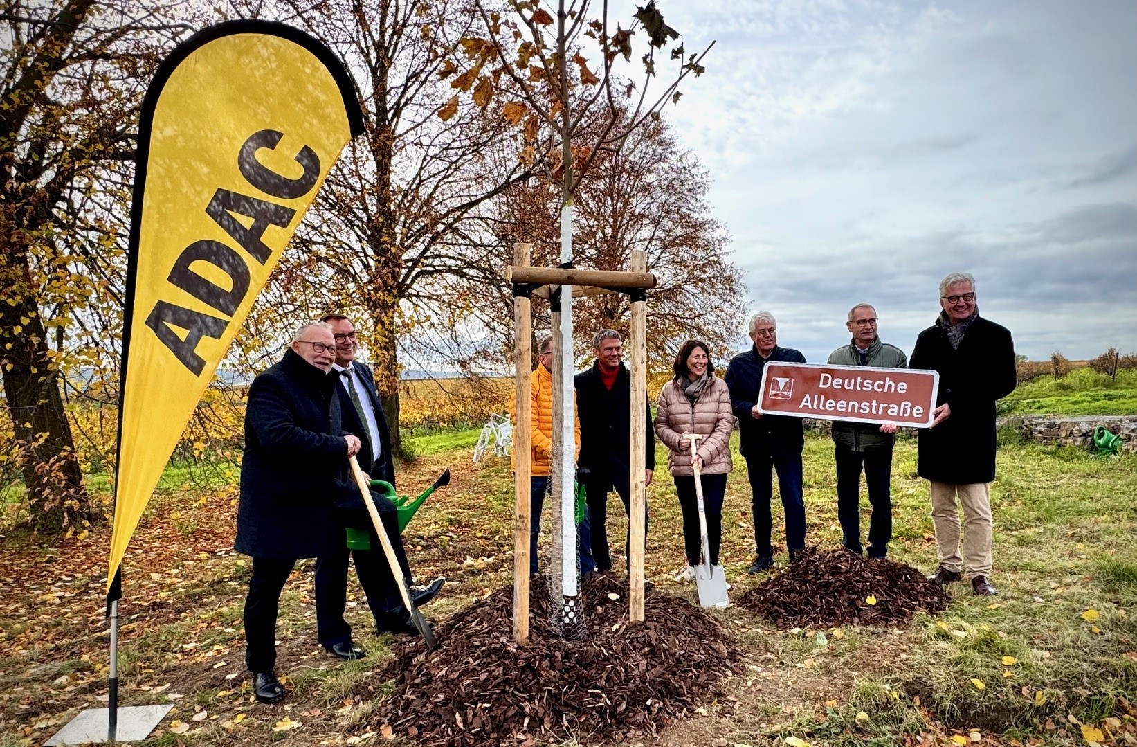 Mehrere Personen stehen um einen neu gesetzten Baum. Links daneben steht eine gelbe ADAC Flagge. Eine Person trägt ein Schild mit der Aufschrift deutsche Alleenstraße.