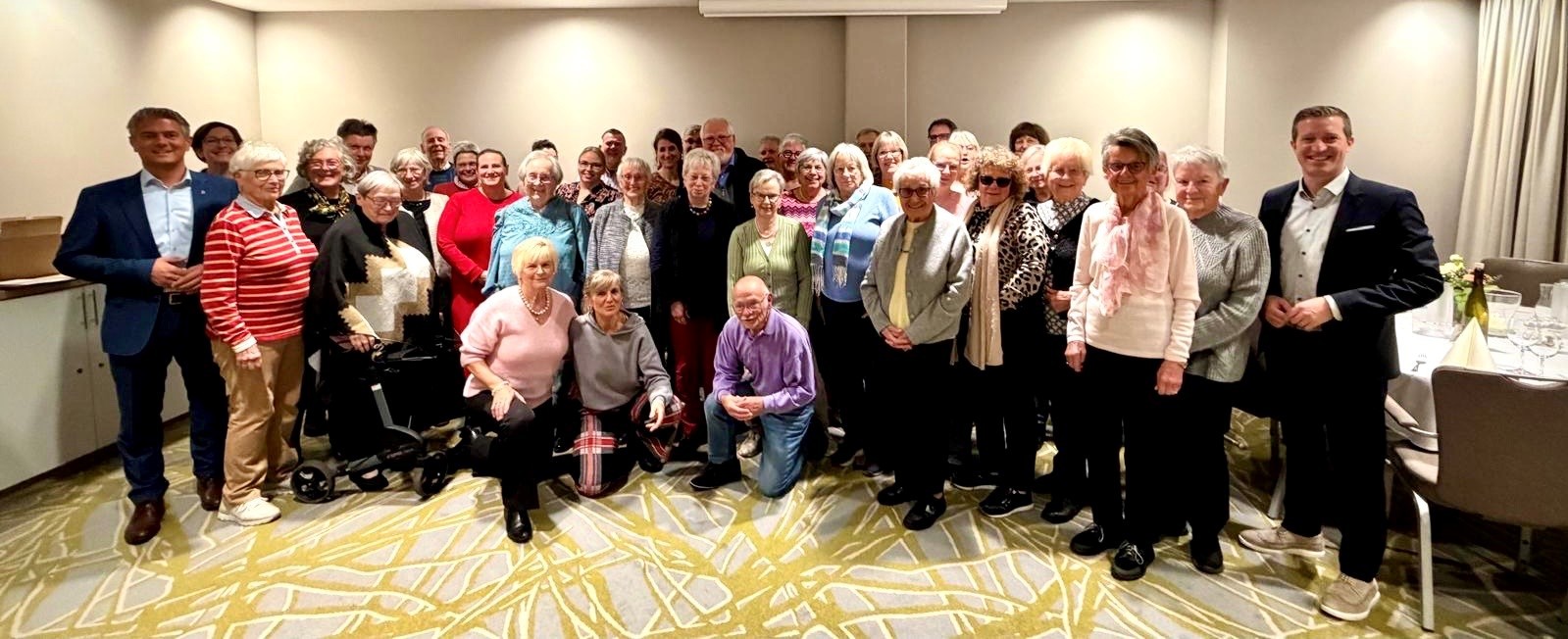 viele ältere Menschen stehen in einem Raum zusammen und schauen in die Kamera