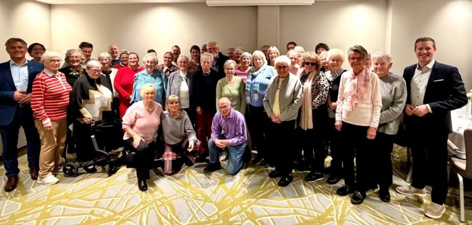 viele ältere Menschen stehen in einem Raum zusammen und schauen in die Kamera