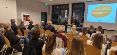 mehrere Personen sitzen in einem Raum und hören einem Vortrag zu