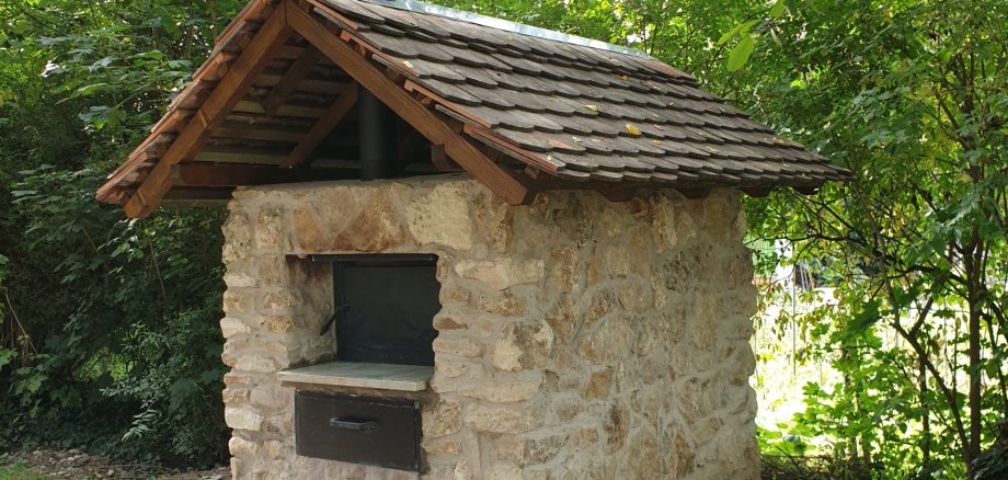 Vor mehreren grünen Bäumen steht ein kleines Steinhäuschen, mit beigenen Steinen und einem roten Ziegeldach. Vorne an dem Häuschen ist ein dunkles fach, welches als Einschubmöglichkeit für Brot diente.