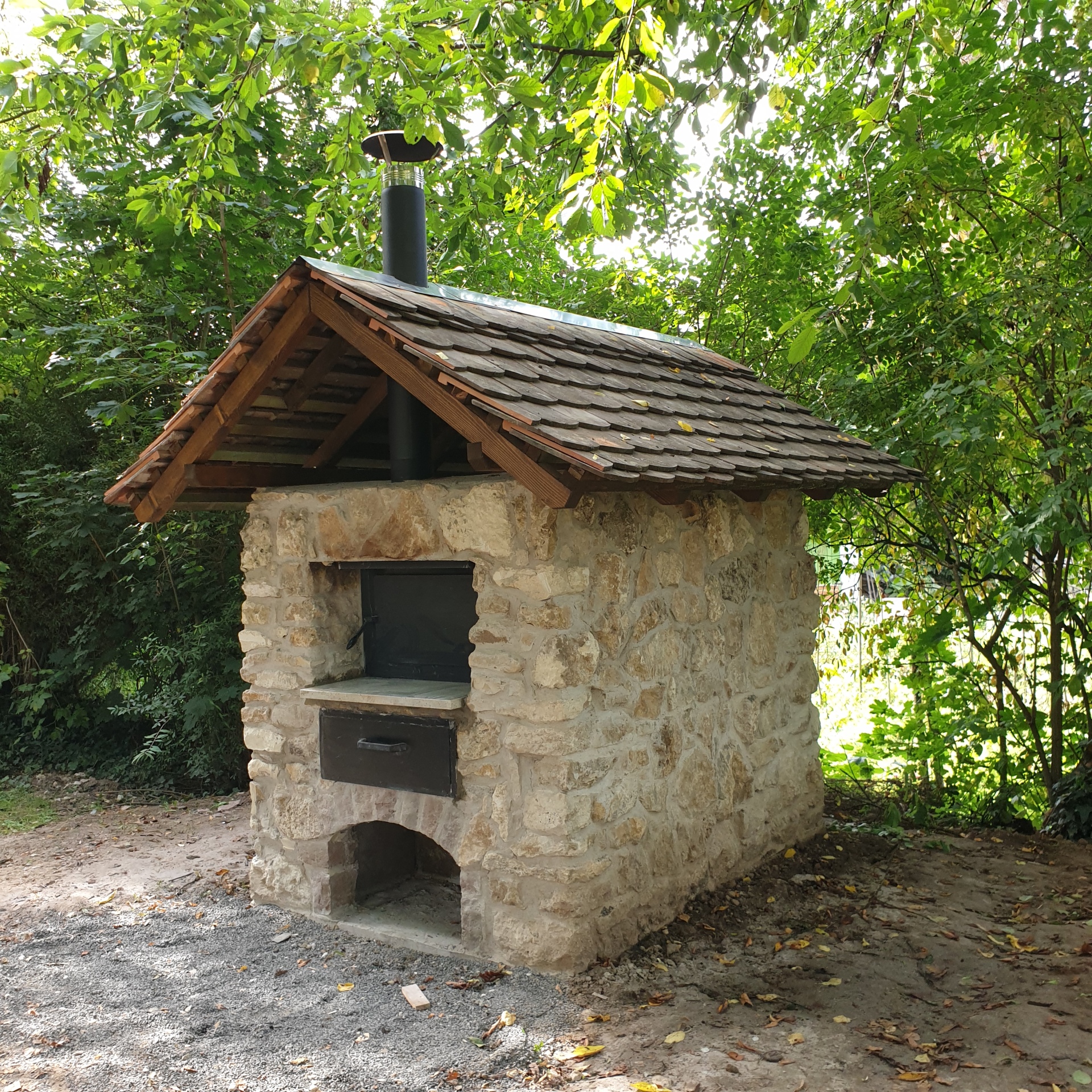 Vor mehreren grünen Bäumen steht ein kleines Steinhäuschen, mit beigenen Steinen und einem roten Ziegeldach. Vorne an dem Häuschen ist ein dunkles fach, welches als Einschubmöglichkeit für Brot diente.