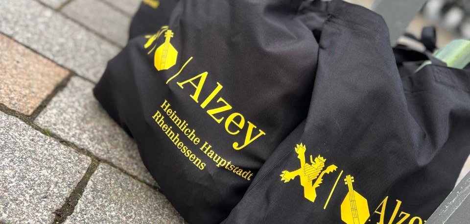 Drei schwarze Alzey Taschen mit dem gelben Stadtlogo stehen befüllt auf dem Boden an einem Stand vom Alzeyer Wochenmarkt.