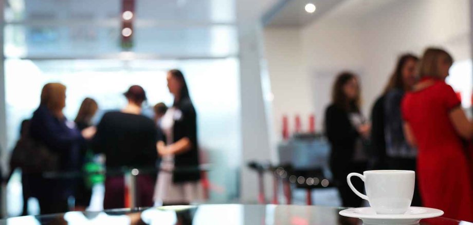 Menschen, die auf einer Veranstaltung sind und im Hintergrund miteinander reden. Im Vordergrund steht eine Tasse Kaffee auf einem Tisch.