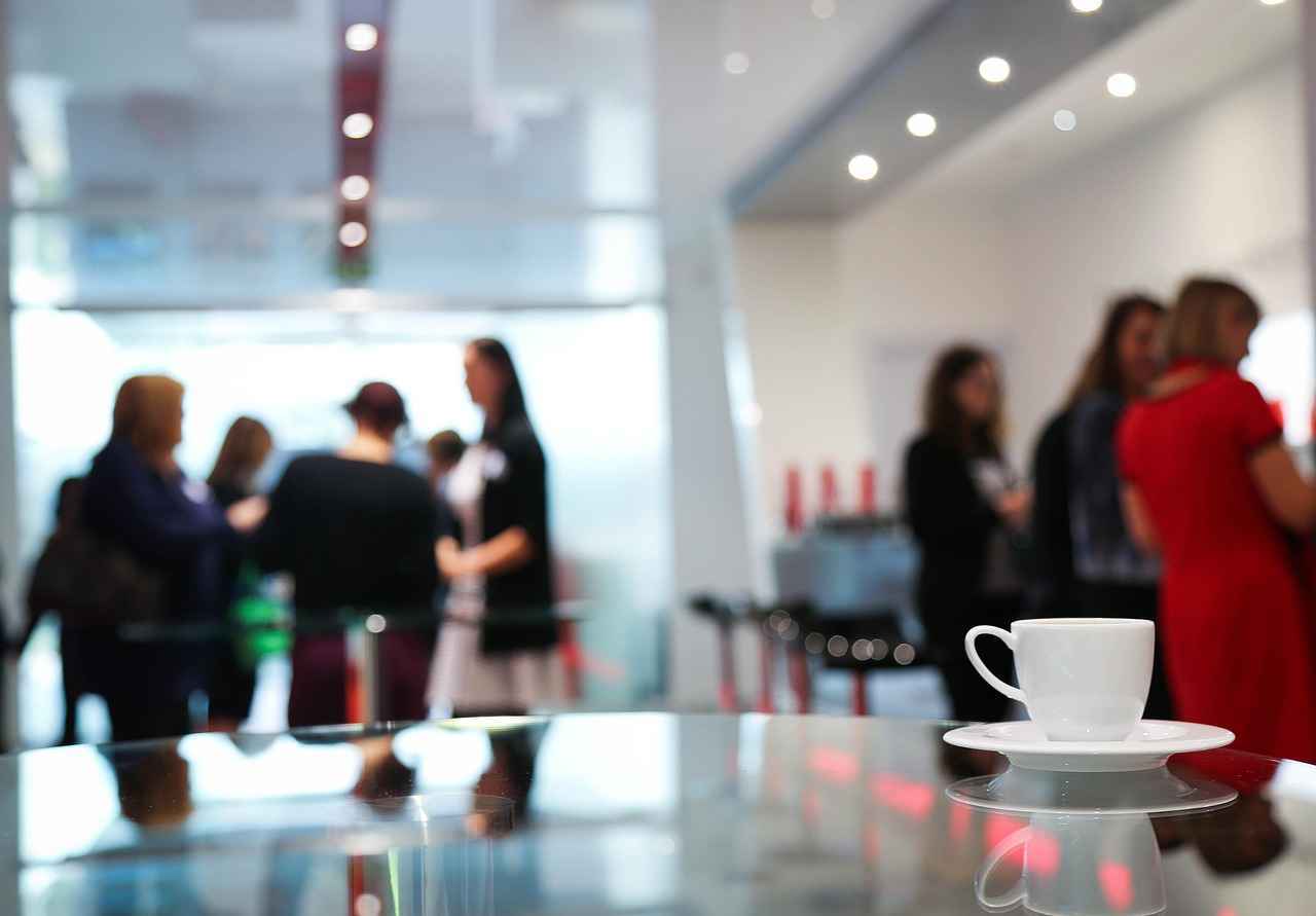 Menschen, die auf einer Veranstaltung sind und im Hintergrund miteinander reden. Im Vordergrund steht eine Tasse Kaffee auf einem Tisch.