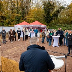 Ein Mann hält auf der Baustelle des ehemaligen Stadtgärtnerei-Geländes eine Rede und einige Mensche, die im Halbkreis vor ihm stehen, hören zu.