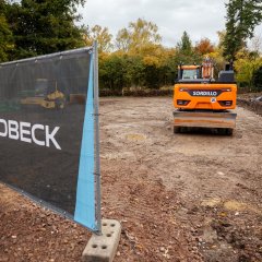 Ein gelber Bagger steht auf dem ehemaligen Stadtgärtnereigelände. Um ihn herum ist nur noch Erde. Im Vordergrund des Bildes ist ein Banner mit der Aufschrift Goldbeck.