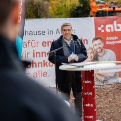 Ein Mann steht vor einem rot-weißen Banner, der die Aufschrift der Alzeyer Baugesellschaft hat. An einem Stehtisch steht ein Mann mit einem blauen Mantel, der etwas in das Mikrofon vor ihm spricht und in die Kamera schaut. Links neben ihm ist eine blaue Fahne mit der Aufschrift Goldbeck aufgestellt.