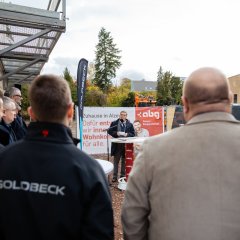 Ein Mann steht vor einem rot-weißen Banner, der die Aufschrift der Alzeyer Baugesellschaft hat. An einem Stehtisch steht ein Mann mit einem blauen Mantel, der etwas in das Mikrofon vor ihm spricht und in die Kamera schaut. Links neben ihm ist eine blaue Fahne mit der Aufschrift Goldbeck aufgestellt. Im Vordergrund des Bildes stehen sechs Männer, die teilweise eine blaue Jacke mit der Aufschrift Goldbeck tragen.