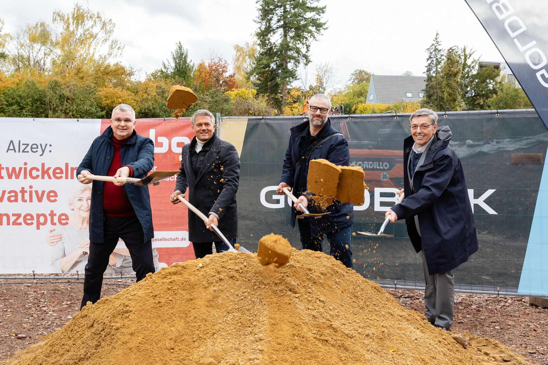 Auf der Baustelle des ehemaligen Stadt-Gärtnereigeländes liegt in der Mitte des Bildes ein hellerer Erdhügel, in dem viel Schaufeln stecken. Dahinter stehen viel Männer, bei denen auch Bürgermeister Steffen Jung dabei ist. Die vier Männer stehen sich im Kreis gegenüber und führen ein Gespräch. Dahinter sind zwei Banner aufgestellt, einer in blau mit der Aufschrift Goldbeck und einer in weiß-rot mit der Aufschrift Alzeyer Baugesellschaft.