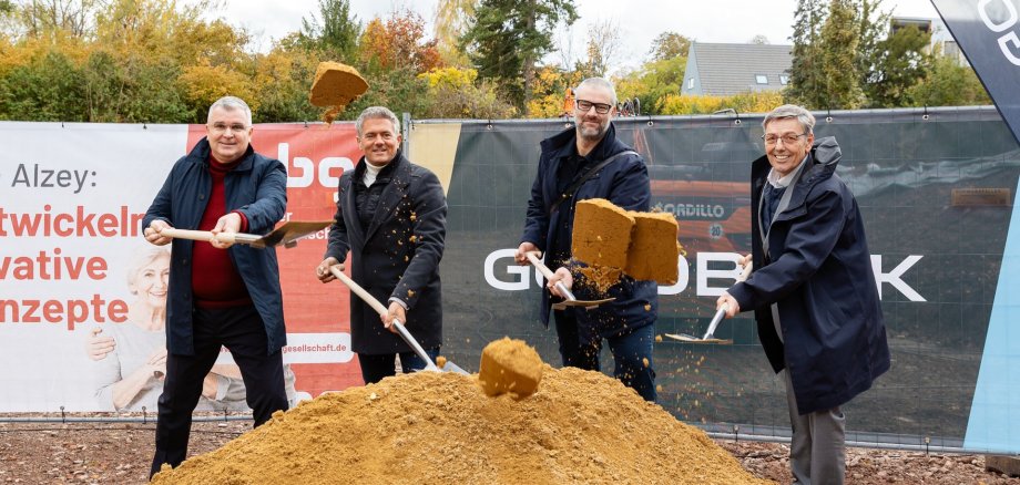 Auf der Baustelle des ehemaligen Stadt-Gärtnereigeländes liegt in der Mitte des Bildes ein hellerer Erdhügel, in dem viel Schaufeln stecken. Dahinter stehen viel Männer, bei denen auch Bürgermeister Steffen Jung dabei ist. Die vier Männer stehen sich im Kreis gegenüber und führen ein Gespräch. Dahinter sind zwei Banner aufgestellt, einer in blau mit der Aufschrift Goldbeck und einer in weiß-rot mit der Aufschrift Alzeyer Baugesellschaft.