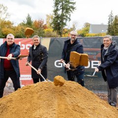 Auf der Baustelle des ehemaligen Stadt-Gärtnereigeländes liegt in der Mitte des Bildes ein hellerer Erdhügel, in dem viel Schaufeln stecken. Dahinter stehen viel Männer, bei denen auch Bürgermeister Steffen Jung dabei ist. Die vier Männer stehen sich im Kreis gegenüber und führen ein Gespräch. Dahinter sind zwei Banner aufgestellt, einer in blau mit der Aufschrift Goldbeck und einer in weiß-rot mit der Aufschrift Alzeyer Baugesellschaft.