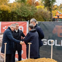 Auf der Baustelle des ehemaligen Stadt-Gärtnereigeländes liegt in der Mitte des Bildes ein hellerer Erdhügel, in dem viel Schaufeln stecken. Dahinter stehen viel Männer, bei denen auch Bürgermeister Steffen Jung dabei ist. Die vier Männer stehen sich im Kreis gegenüber und führen ein Gespräch. Dahinter sind zwei Banner aufgestellt, einer in blau mit der Aufschrift Goldbeck und einer in weiß-rot mit der Aufschrift Alzeyer Baugesellschaft.