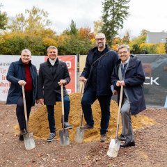 Auf der Baustelle des ehemaligen Stadt-Gärtnereigeländes liegen Berge von Erde in der rechten Hälfte des Bildes. In der Mitte liegt ein hellerer Erdhügel, vor dem vier Männer stehen. Einer von ihnen ist Bürgermeister Steffen Jung. Alle haben eine Schaufel in der Hand. auf der sie mit einem Fuß stehen. Dahinter sind zwei Banner aufgestellt, einer in blau mit der Aufschrift Goldbeck und einer in weiß-rot mit der Aufschrift Alzeyer Baugesellschaft. Hinter den Bannern stehen zwei Bagger, einer in gelb und einer in organge.