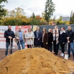 Auf der Baustelle des ehemaligen Stadt-Gärtnereigeländes liegen Berge von Erde in der rechten Hälfte des Bildes. In der Mitte liegt ein hellerer Erdhügel, hinter dem neun Personen stehen, von denen viel Leute eine Schaufel in der Hand tragen. Dahinter sind zwei Banner aufgestellt, einer in blau mit der Aufschrift Goldbeck und einer in weiß-rot mit der Aufschrift Alzeyer Baugesellschaft.