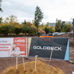 Auf der Baustelle des ehemaligen Stadt-Gärtnereigeländes liegen Berge von Erde in der rechten Hälfte des Bildes. In der Mitte liegt ein hellerer Erdhügel, in dem vier Schaufeln stecken. Dahinter sind zwei Banner aufgestellt, einer in blau mit der Aufschrift Goldbeck und einer in weiß-rot mit der Aufschrift Alzeyer Baugesellschaft. Im Hintergrund stehen zwei Bagger, ein gelber und ein organgener.