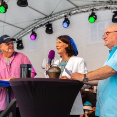 Auf dem Bild sind drei Menschen zu sehen, die nebeneinander an einem Stehtisch auf einer Bühne stehen. Ihr Blick ist Richtung Publikum gerichtet und Daniela Schmitt in der Mitte, hat ein Glas Wein in der Hand und hält in der anderen ein Mikrofon, durch das sie dem Publikum etwas erzählt.
