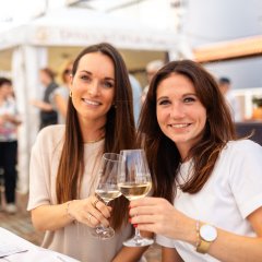 Auf dem Bild sind zwei Frauen zu sehen, die beide in die Kamera schauen und mit einem Glas Wein anstoßen.
