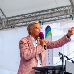 Auf dem Bild sieht man einen Mann, der auf einer Bühne steht und etwas in das Mikrofon sagt. Er schaut in Richtung Zuschauer und gestikuliert mit seiner linken Hand.