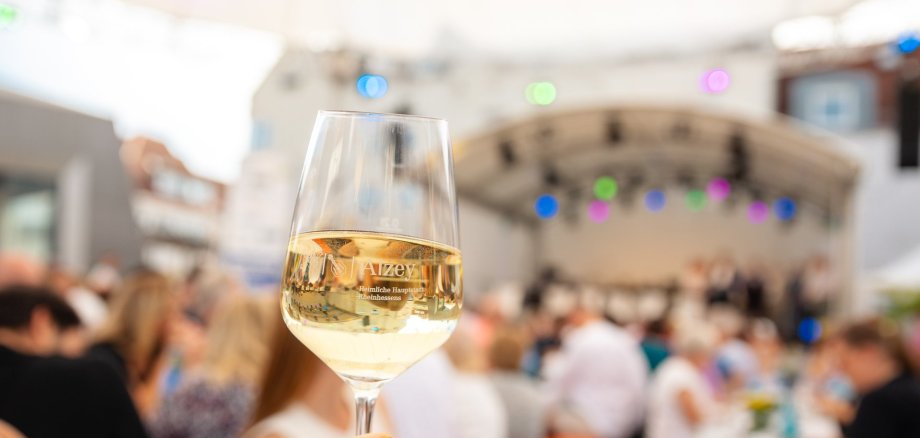 Auf dem Foto ist ein Weinglas, befüllt mit Wein zu sehen, welches in die Kamera gehalten wird. Im Hintergrund sieht man verschwommen die Besucher der Weinprobe vor der Bühne sitzen.