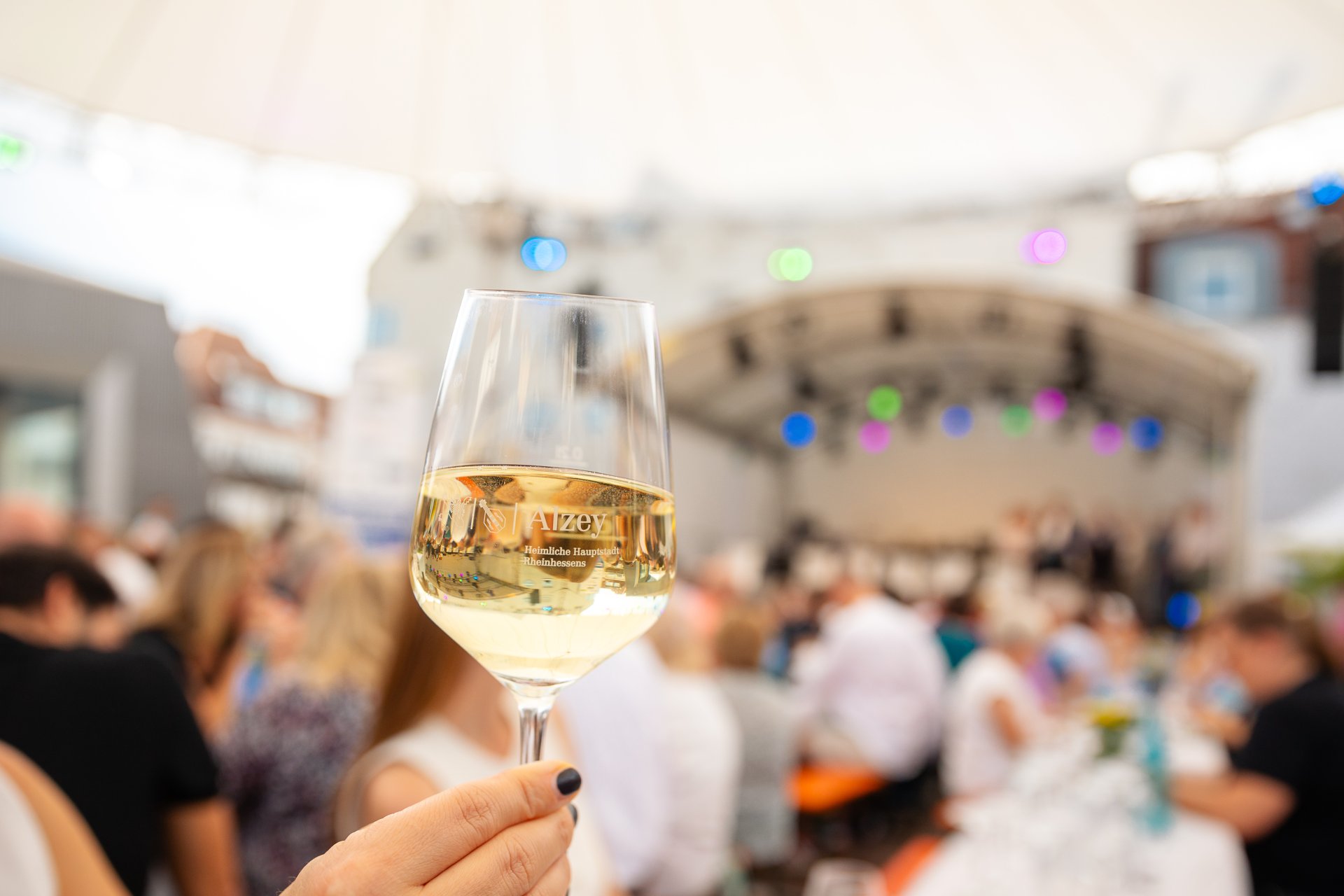 Auf dem Foto ist ein Weinglas, befüllt mit Wein zu sehen, welches in die Kamera gehalten wird. Im Hintergrund sieht man verschwommen die Besucher der Weinprobe vor der Bühne sitzen.