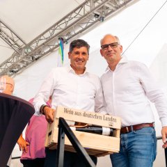 Auf dem Bild sieht man zwei Männer, die grade dabei sind von der Bühne zu gehen. Der eine hat eine kleine hölzerne Weinkiste, befüllt mit Weinflaschen in der Hand.