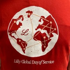 Auf dem Bild ist ein rotes T-Shirt zu erkennen, was eine weiße Weltkugel als Aufdruck hat. Auf dieser Weltkarte sind überall verteilt Punkte eingezeichnet, die durch Linien vernetzt sind. Dazwischen ist ein großes geschwungenes L in rot zu erkennen.