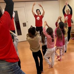 Auf dem Bild sieht man drei erwachsene Personen, die zusammen mit mehreren Kindern zusammen eine Tanzübung im Stehen machen.