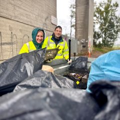 Auf dem Bild sind zwei Personen zu sehen, die in die Kamera lachen. Sie tragen gelbe Warnwesten und stehen vor einem Container, in den sie gesammelten Müll werfen.
