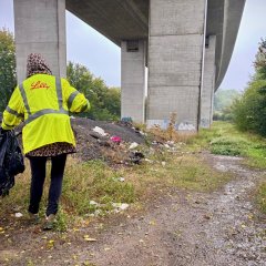 Eine Person mit einer neon gelben Warnjacke läuft auf einem Faldweg unter einer Autobahnbrücke lan. Sie hält in der linken Hand einen Müllsack und sammelt mit einer Zange in ihrer rechten Hand Müll auf.