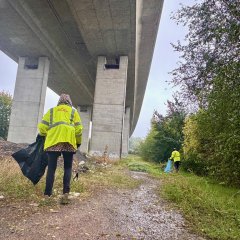 Eine Person mit einer neon gelben Warnjacke läuft auf einem Faldweg unter einer Autobahnbrücke lan. Sie hält in der linken Hand einen Müllsack und sammelt mit einer Zange in ihrer rechten Hand Müll auf.