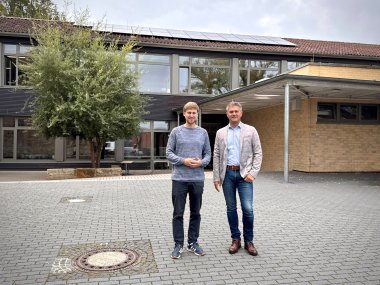 Zu sehen sind auf dem Bild zwei Männer im Vordergrund. Im Hintergrund sieht man neben einem Baum auch eine Schule.
